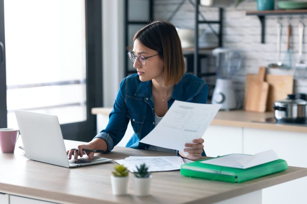 young woman working with computer while consulting some invoices and documents in the kitchen